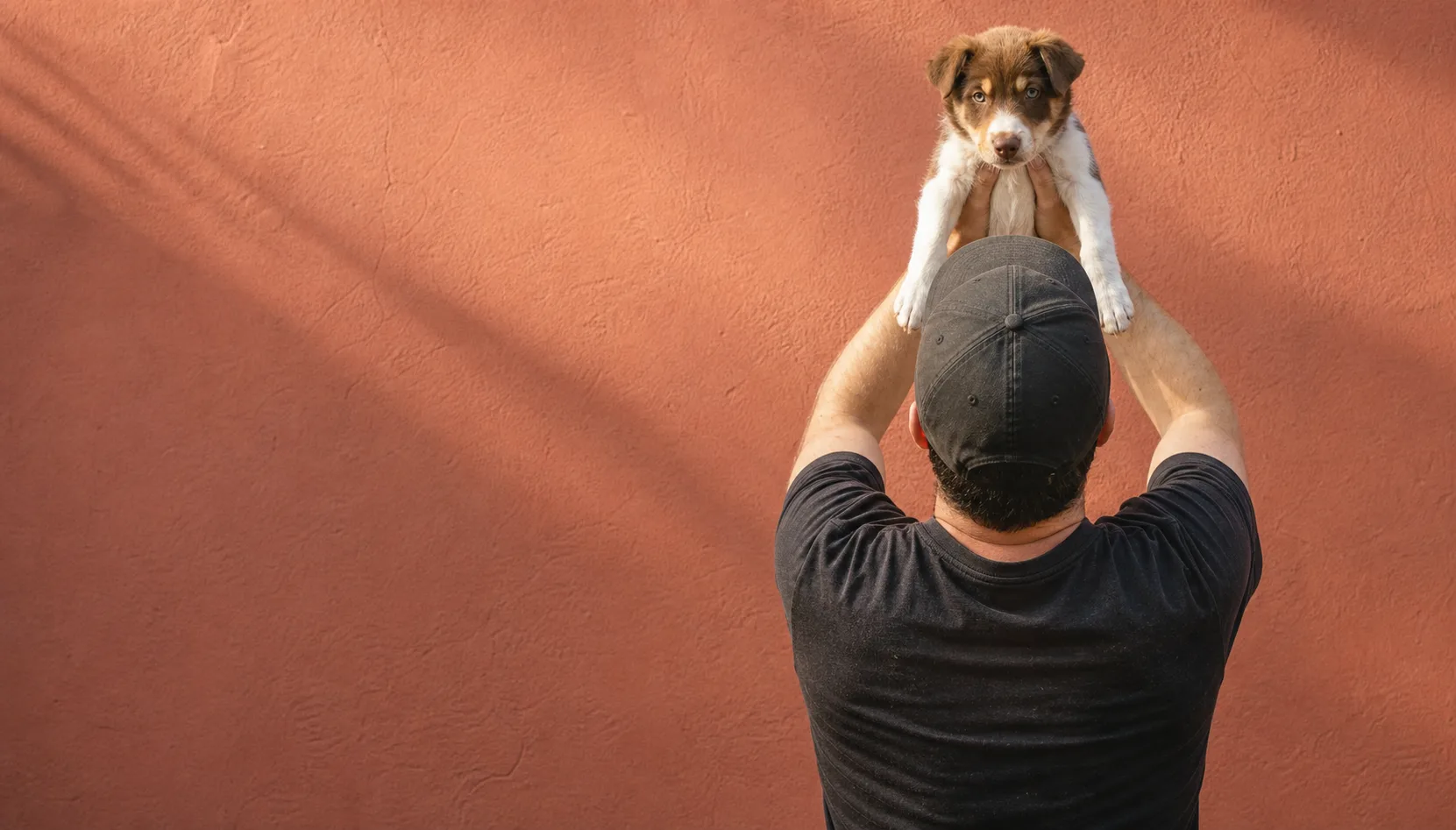 Adiestrador de espaldas levantando un cachorro frente a una pared terracota
